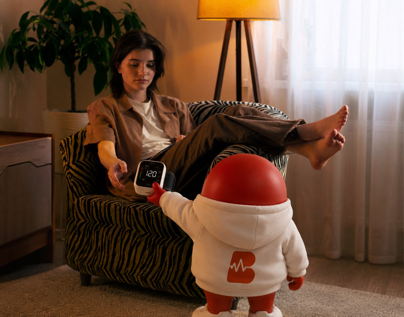 Woman relaxing on a couch while the red Blood Health mascot hands her the wrist blood pressure monitor